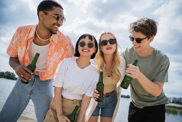 young multicultural friends smiling while holding beer on beach - Foto, afbeelding