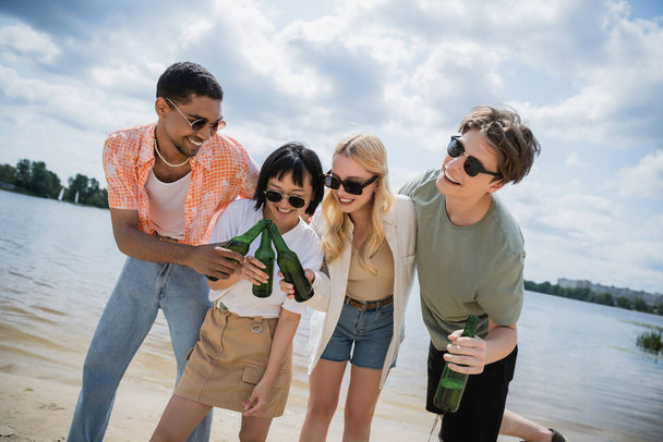 low angle view of multicultural friends in sunglasses clinking refreshing beer on beach - Photo, image