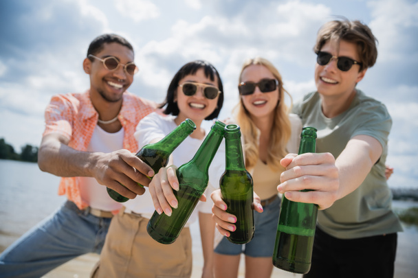 межрасовые друзья в солнечных очках, звенящие пивные бутылки на размытом фоне - Фото, изображение