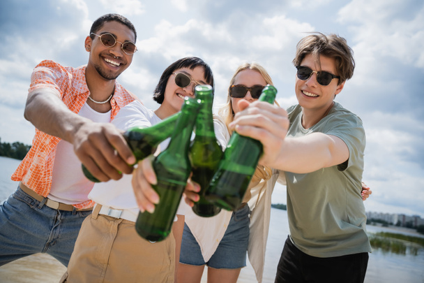 joyful multicultural friends toasting with beer bottles on riverside - Photo, image