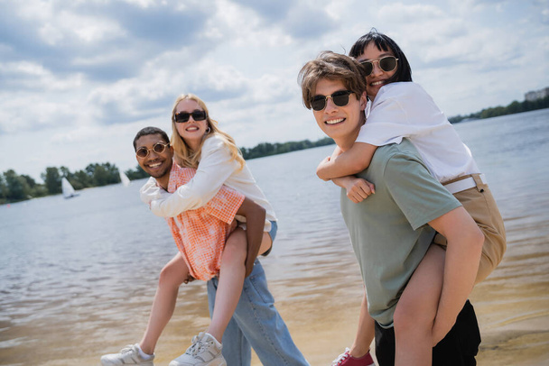young multicultural men in sunglasses looking at camera while piggybacking women near river - Foto, Imagen