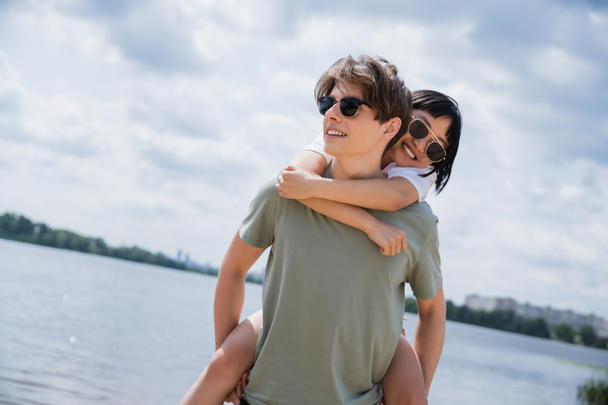 young man in sunglasses piggybacking cheerful asian woman near river - Foto, Imagen