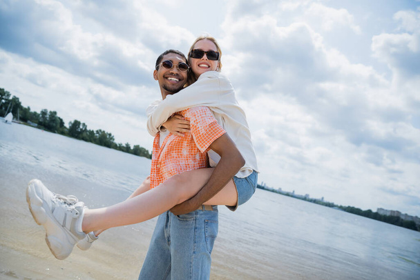 happy african american man piggybacking smiling girlfriend near river - Фото, изображение