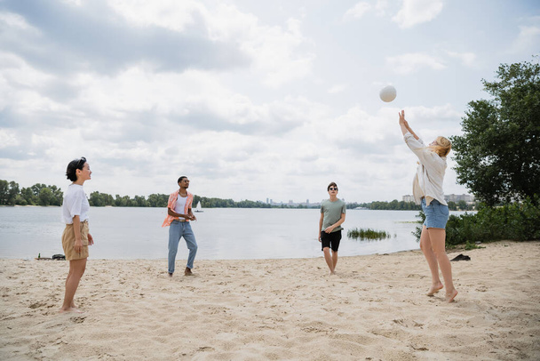 young woman passing ball to multiethnic friends playing beach volleyball on riverside - Fotó, kép