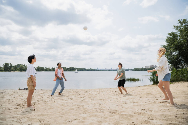 молодые многонациональные друзья играют в волейбол на песчаном пляже у реки - Фото, изображение