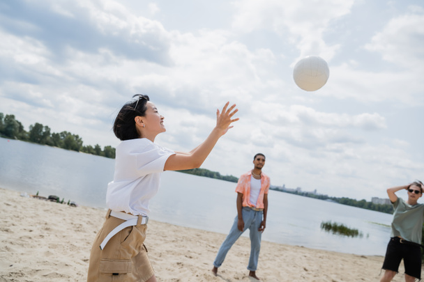 возбужденная азиатка, играющая в волейбол с межрасовыми мужчинами на песчаном пляже - Фото, изображение
