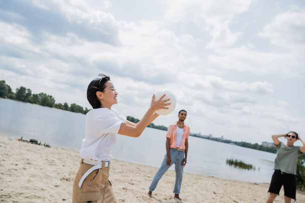 χαρούμενα ασιατική γυναίκα κρατώντας μπάλα, ενώ παίζει βόλεϊ στην παραλία με πολυεθνικούς φίλους - Φωτογραφία, εικόνα