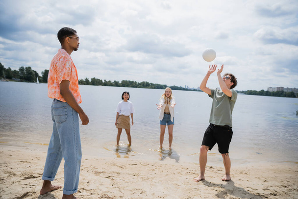 young man passing ball while playing beach volleyball with interracial friends - Fotó, kép
