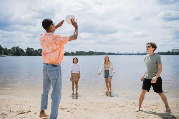 african american man passing ball while playing beach volleyball with multiethnic friends - Fotó, kép