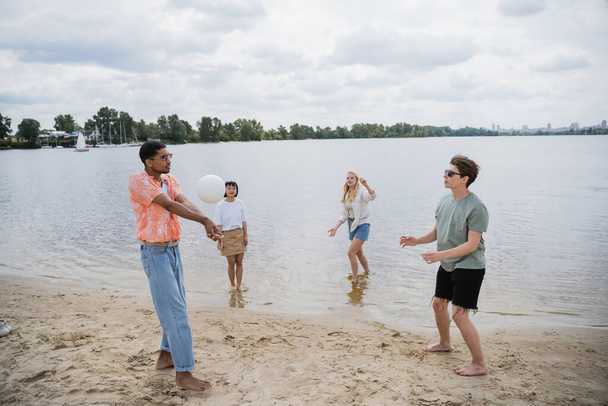 διαφυλετικοί άνδρες που παίζουν beach volley κοντά σε γυναίκες που στέκονται στο νερό - Φωτογραφία, εικόνα