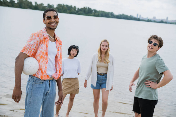 african american man with ball smiling at camera near multiethnic friends on beach - Photo, Image