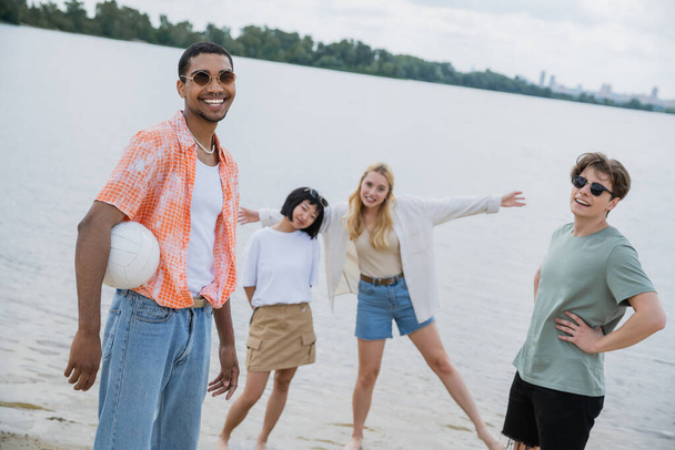 young african american man in sunglasses holding ball and looking at camera near multiethnic friends - 写真・画像