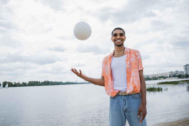 ευτυχισμένος Αφροαμερικάνος με γυαλιά ηλίου κοιτάζοντας κάμερα ενώ παίζει με μπάλα κοντά στο ποτάμι - Φωτογραφία, εικόνα