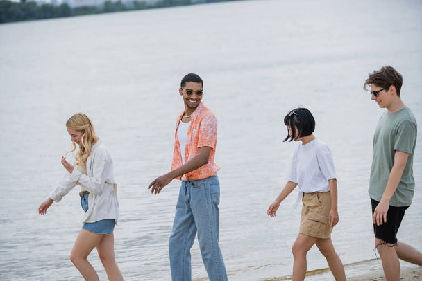 cheerful african american man in sunglasses walking with interracial friends near river - 写真・画像