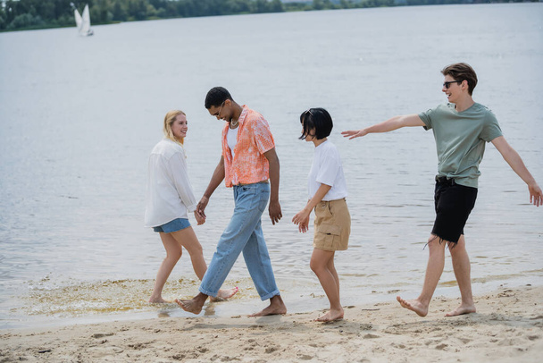 excited man walking with multiethnic friends near river and pointing with hand - Photo, image