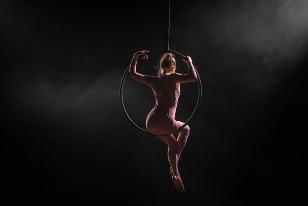 A young woman aerialist sitting on an aerial ring under a circus dome. Athlete acrobat performing in the dark with backlight and smoke. View from the back of a slender flexible female equilibrist. For - Foto, Bild