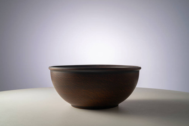 Empty earthen deep bowl on white table isolated on gray studio background. Brown handicraft ramekin or rustic plate. Empty earthenware, crockery from clay for food. Old dishes round shape - Photo, image