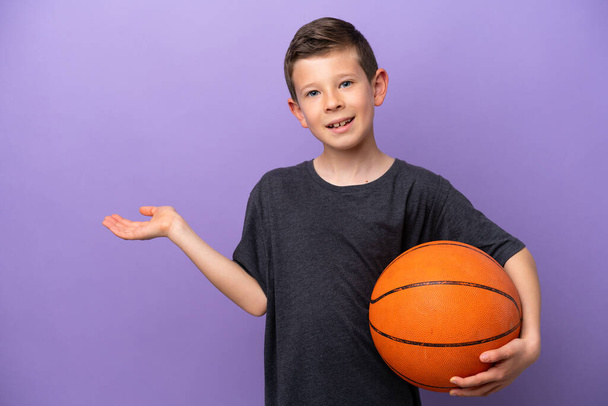 Little boy playing basketball isolated on purple background extending hands to the side for inviting to come - Φωτογραφία, εικόνα