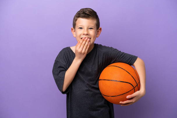Little boy playing basketball isolated on purple background happy and smiling covering mouth with hand - 写真・画像