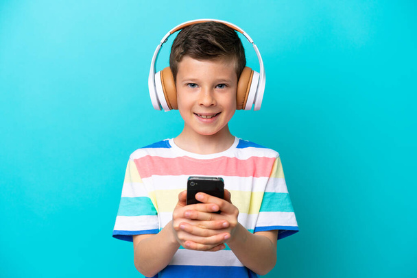 Μικρό αγόρι απομονωμένο σε μπλε φόντο ακούγοντας μουσική με ένα κινητό και κοιτάζοντας μπροστά - Φωτογραφία, εικόνα