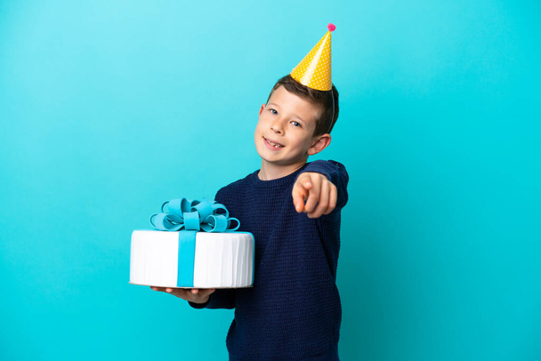 Маленький хлопчик тримає торт на день народження ізольований на синьому фоні, вказуючи спереду з щасливим виразом
 - Фото, зображення