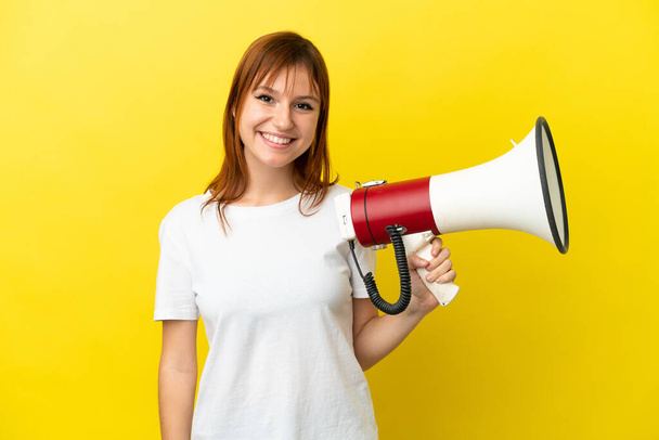 Redhead girl isolated on yellow background holding a megaphone and smiling a lot - Fotografie, Obrázek