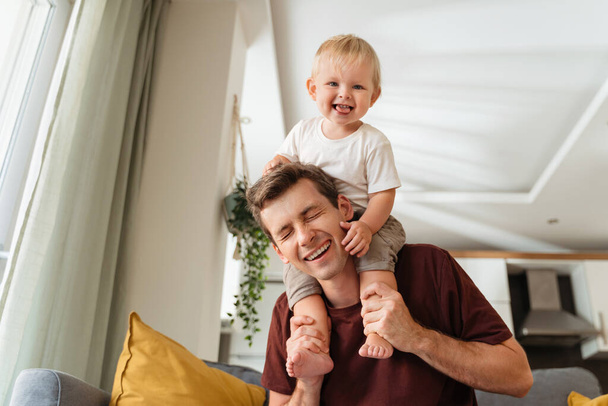 Laughing father giving his kid piggyback ride on shoulders, baby boy pulling daddys hair, father and son spending time together on sunday morning in living-room. Dad playing with his toddler boy - 写真・画像