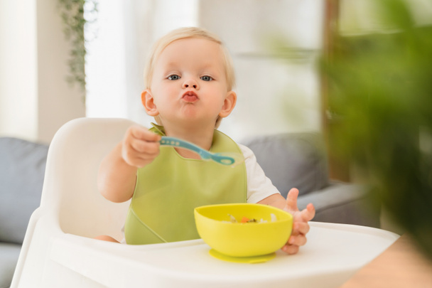 Adorable blond baby boy sitting at table in high chair with spoon in hand and yellow plate with breakfast in front of him, training skills to feed himself, wearing green bib. Baby eating by himself - 写真・画像