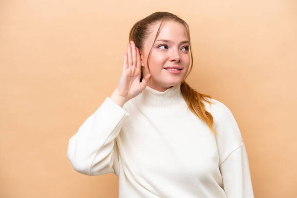 Молода біла жінка ізольована на бежевому фоні, слухаючи щось, поклавши руку на вухо
 - Фото, зображення