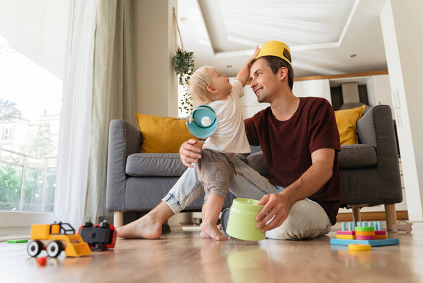 Молодий батько і син розважаються разом. Блондинка малюк кладе іграшку на голову татам, сидячи на підлозі у вітальні, тато розважає свого маленького хлопчика, грає в іграшки, активні ігри вдома
 - Фото, зображення