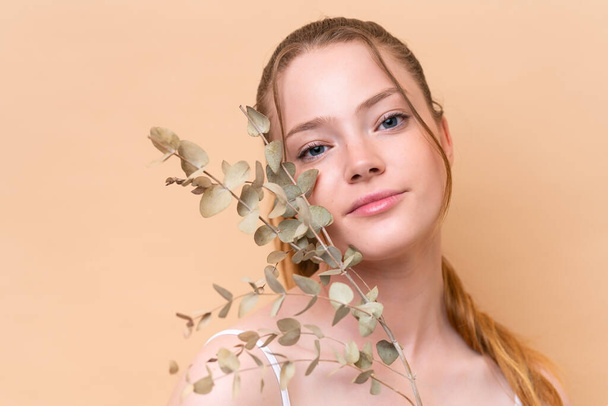 Joven chica caucásica aislada sobre fondo beige sosteniendo una rama de eucalipto. Primer plano retrato - Foto, imagen