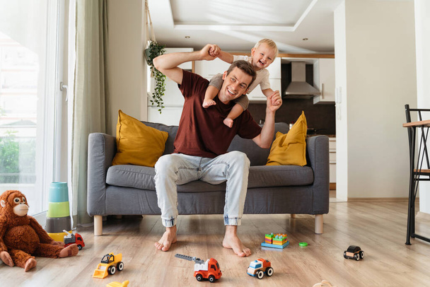 Счастливый отец, несущий смеющегося маленького блондинчика на шее и сидящий на диване в гостиной, окруженный игрушками, проводит вместе выходные. Молодой отец играет со своим малышом - Фото, изображение