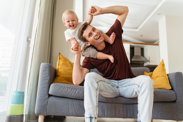Father and son relationships. Handsome dad carrying baby boy on his shoulders, bending to side pretending to fall making cute kid laugh with excitement sitting on grey sofa. Fatherhood, parenting - Foto, imagen