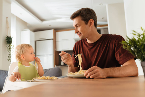 Вид сбоку на счастливого отца и маленького мальчика, сидящего за кухонным столом со спагетти, сына в слюнявчике в стульчике, который учится есть вилкой. Самокормление, отлучение - Фото, изображение