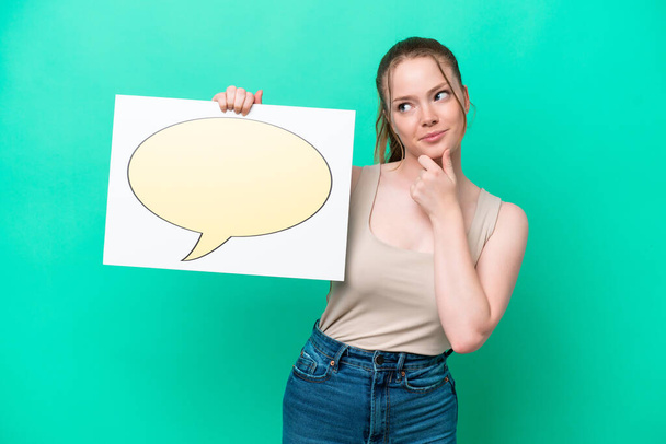 Young caucasian woman isolated on green background holding a placard with speech bubble icon and thinking - Fotó, kép
