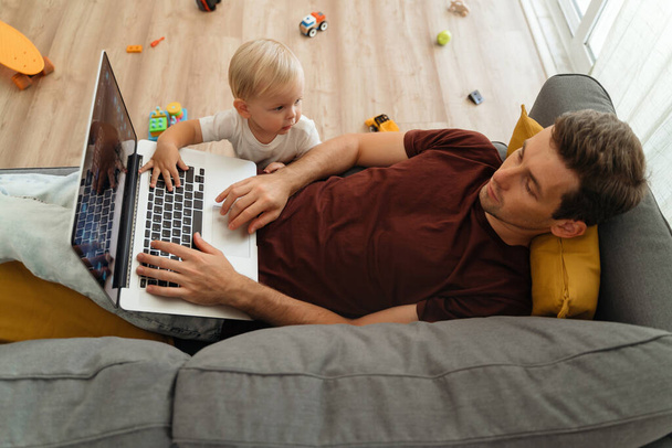Naughty little baby boy bothering his father lying on couch with laptop in living-room, tapping keyboard with hand, disturbing working from home or resting dad. Toddler son asking for dad attention - 写真・画像