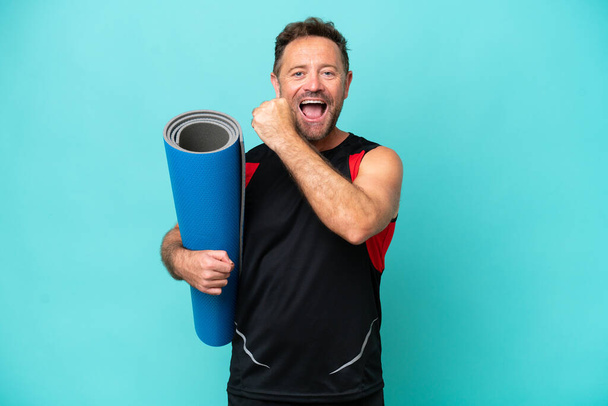 Middle age sport man going to yoga classes while holding a mat isolated on blue background celebrating a victory - Photo, Image