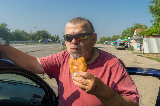 Portrait of hungry Ukrainian senior driver taking patty near his car while standing near Vasylivka, Zaporizhzhia Oblast, Ukraine - Zdjęcie, obraz