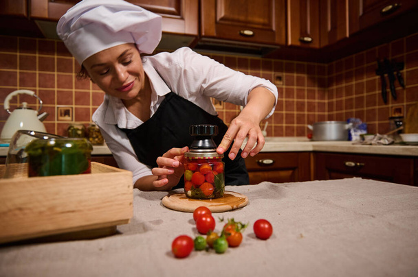 Beautiful Hispanic housewife in white chefs cap and black apron, using seamer, closes lid of jar with ripe and juicy tomatoes while canning according to traditional family recipe in home kitchen - Foto, afbeelding