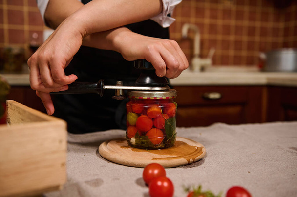 Process of preserving cherry tomatoes for the winter. Womens hands close the lids of jars with ripe red juicy organic tomatoes with a special key. Canning food according traditional family recipe - Foto, immagini