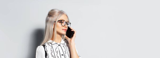 Studio portrait of pretty smiling girl with blonde hair, talking on smartphone, wearing eyeglasses on white background, panoramic banner view with copy space. - 写真・画像