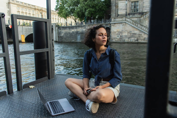 surprised freelancer holding smartphone while sitting with crossed legs near laptop and river in berlin  - 写真・画像