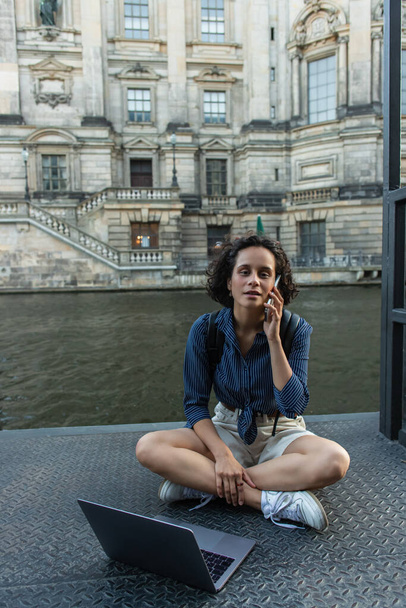σγουρή γυναίκα μιλάει στο smartphone, ενώ κάθεται με σταυρωμένα πόδια κοντά στο laptop και το ποτάμι στο Βερολίνο  - Φωτογραφία, εικόνα