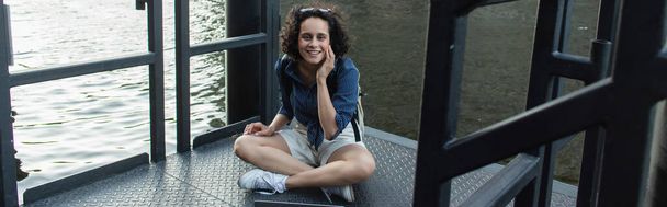 σγουρά νεαρή γυναίκα χαμογελά και κάθεται με σταυρωμένα πόδια κοντά στο ποτάμι στο Βερολίνο, πανό - Φωτογραφία, εικόνα