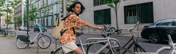 περίεργη νεαρή γυναίκα με καλοκαιρινά ρούχα στέκεται κοντά στο ποδήλατο στο Βερολίνο, πανό - Φωτογραφία, εικόνα
