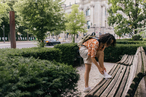 curly young woman in summer outfit tying shoelaces in green park in berlin  - Foto, Imagen