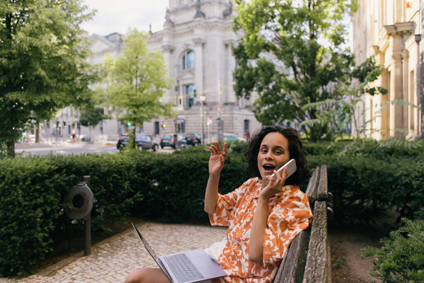 здивований молодий фрілансер в літньому вбранні сидить на лавці з ноутбуком і розмовляє на смартфоні в зеленому парку
  - Фото, зображення