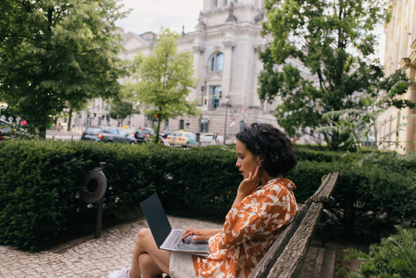 вид збоку молода кучерява жінка сидить на лавці і використовує ноутбук у зеленому парку
  - Фото, зображення