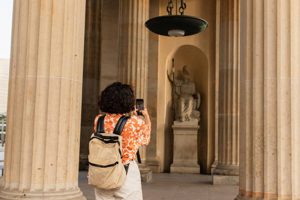 back view of curly woman with backpack taking photo of statue on smartphone  - Photo, Image