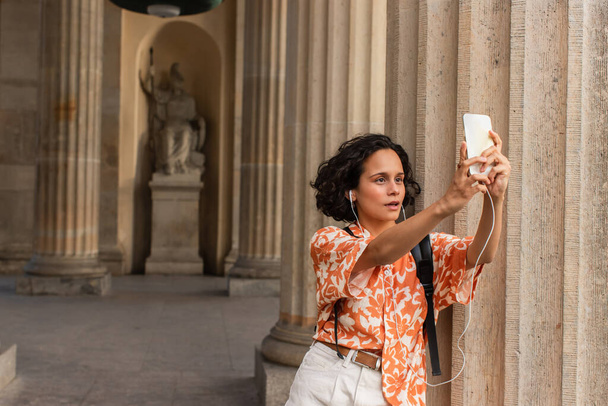 curly young tourist in wired earphones taking selfie with statue while holding smartphone  - Zdjęcie, obraz